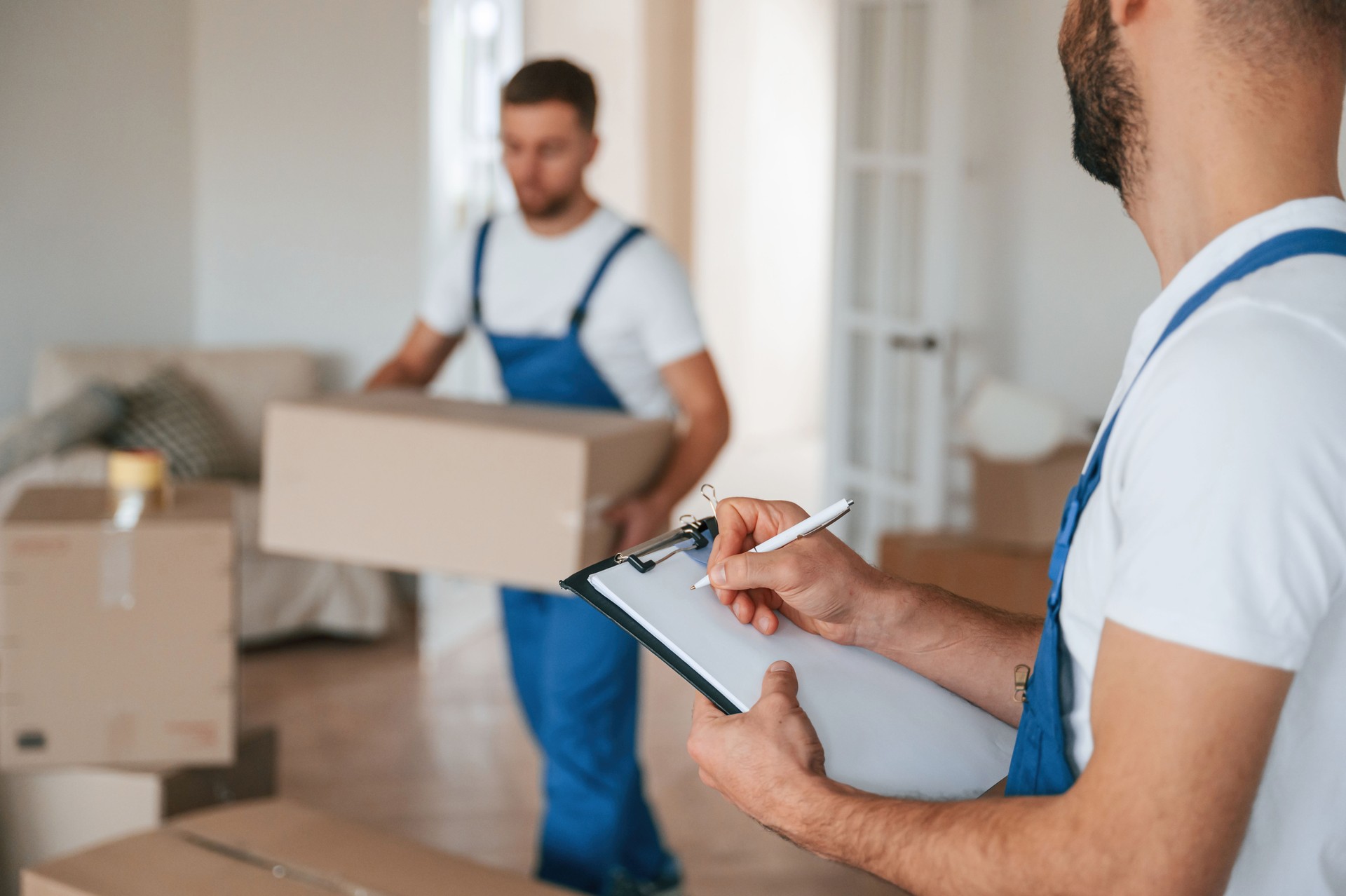 Writing in notepad, holding box. Two moving service employees in a room